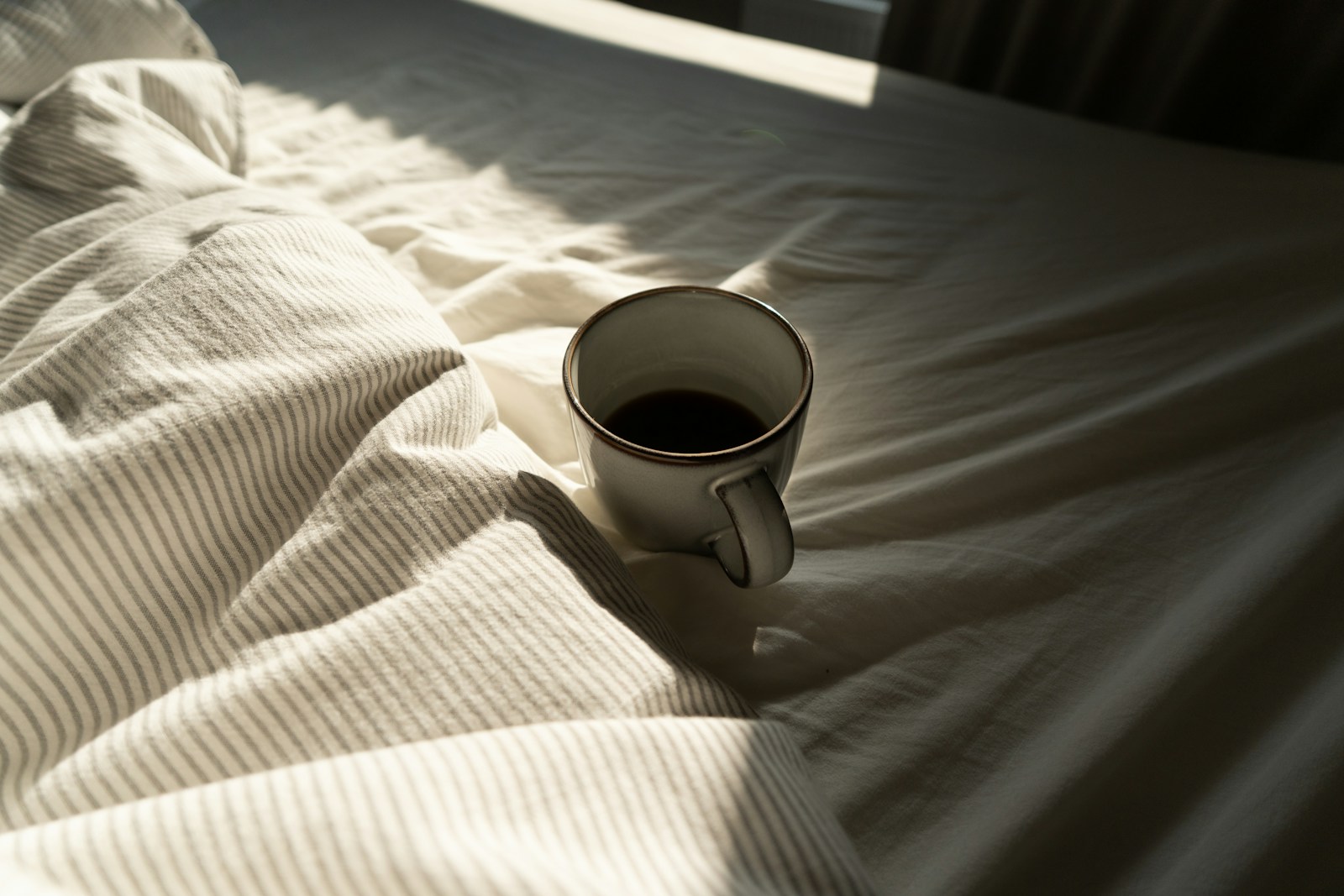 A mug of coffee resting on rumpled white bedding in soft morning light
